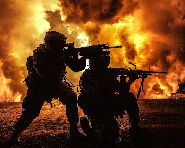 Silhouettes de deux soldats de l'armée, équipe de marines américains en action, entouré de feu et de fumée, tirant avec un fusil d'assaut et une mitrailleuse, attaquant l'ennemi avec des coups de feu suppressifs pendant une mission offensive