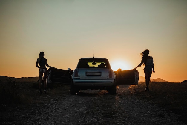 Silhouettes féminines sur fond de coucher de soleil à côté de la voiture avec portes ouvertes