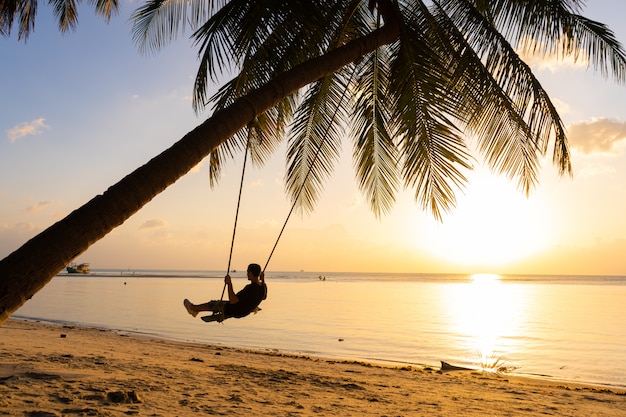 Silhouettes d'un gars sur une balançoire accrochée à un palmier, regardant le coucher du soleil dans l'eau.