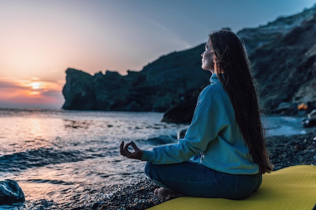 Silhoute de jeune femme aux cheveux longs en jeans et vêtements de sport pratiquant à l'extérieur sur un tapis de yoga