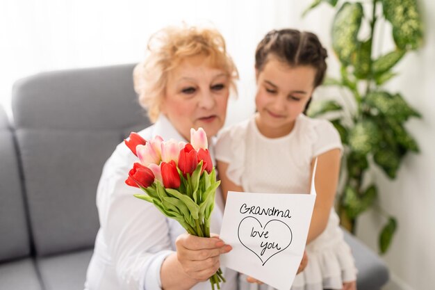 Sincère mignonne petite-fille forte câlinant et embrassant dans la joue sa grand-mère des années 60 lui a donné de jolies fleurs de printemps félicite avec anniversaire, Journée internationale de la femme, image de concept en gros plan.