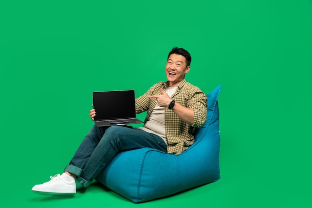 Site Web cool heureux homme asiatique mature pointant sur un ordinateur portable avec écran blanc assis sur une chaise pouf