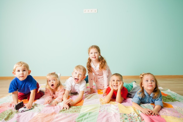 Six enfants adorables et excités allongés sur un sol sur fond bleu. Les enfants regardent quelque chose d'intéressant derrière la caméra avec enthousiasme et intérêt sur leurs visages. Concept d'école à la maison