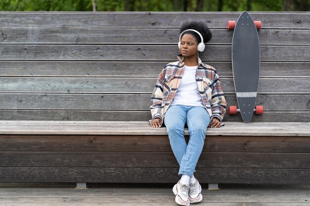 Skateboarder femme noire détendue écouter de la musique s'asseoir dans le parc avec concept de mode de vie urbain de long conseil