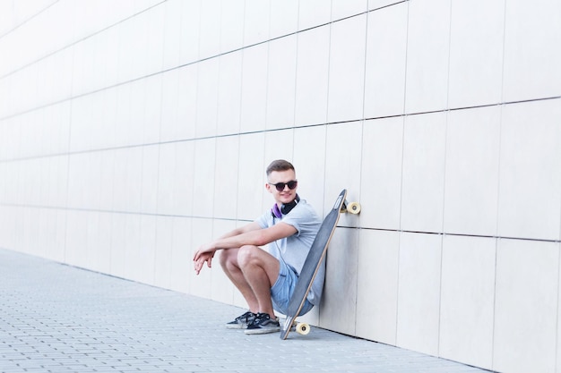 Skateur vêtu d'un t-shirt gris et de lunettes de soleil assis avec un longboard devant un mur de briques de construction moderne