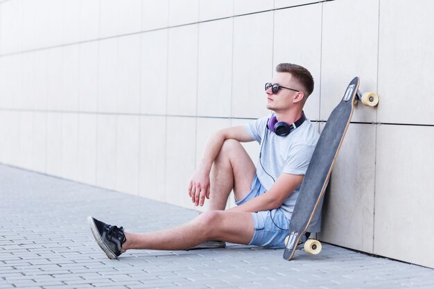 Skateur vêtu d'un t-shirt gris et de lunettes de soleil assis avec un longboard devant un mur de briques de construction moderne