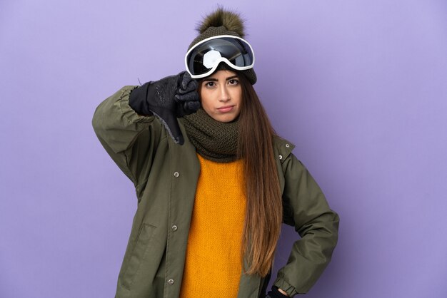 Skieur caucasian girl avec des lunettes de snowboard isolé sur mur violet montrant le pouce vers le bas avec une expression négative