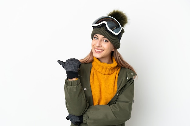 Skieur caucasien femme avec des lunettes de snowboard isolé sur fond blanc pointant vers le côté pour présenter un produit