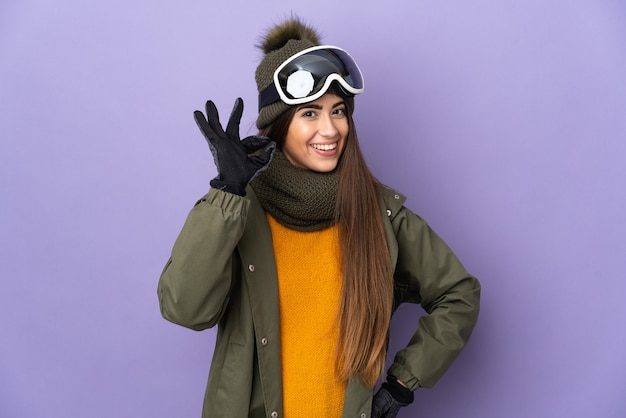 Skieur fille caucasienne avec des lunettes de snowboard isolé sur fond violet montrant signe ok avec les doigts