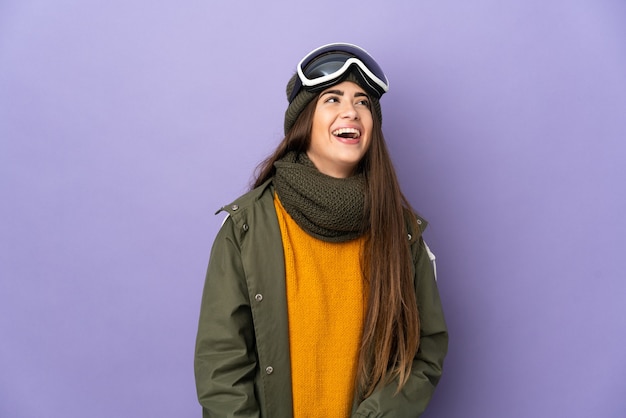 Skieur fille caucasienne avec des lunettes de snowboard isolé sur mur violet en riant