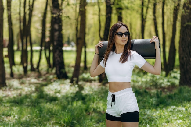 Slim fit jeune femme en tenue de sport tenant un tapis de yoga debout à l'extérieur et regardant loin Attrayante sportive caucasienne par une journée ensoleillée dans le parc Concept de mode de vie actif d'entraînement sportif