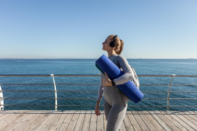 Slim woman fit écouter de la musique avec des écouteurs et tenant un tapis à une journée ensoleillée sur la plage