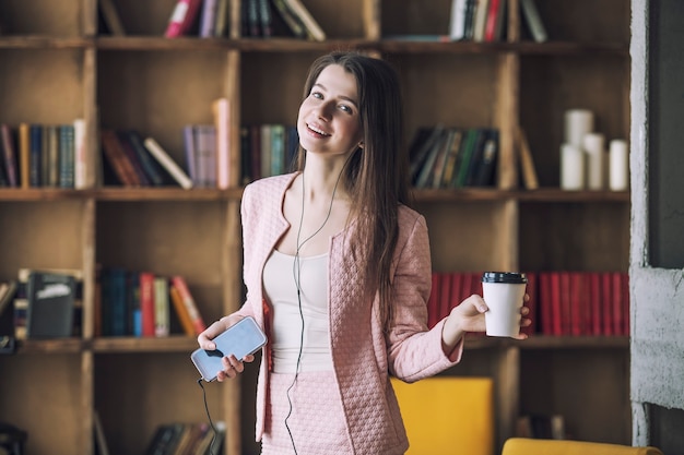 Smart belle jeune femme dans les écouteurs avec un verre de café à la main au lieu d'un microphone dansant et chantant sur la musique