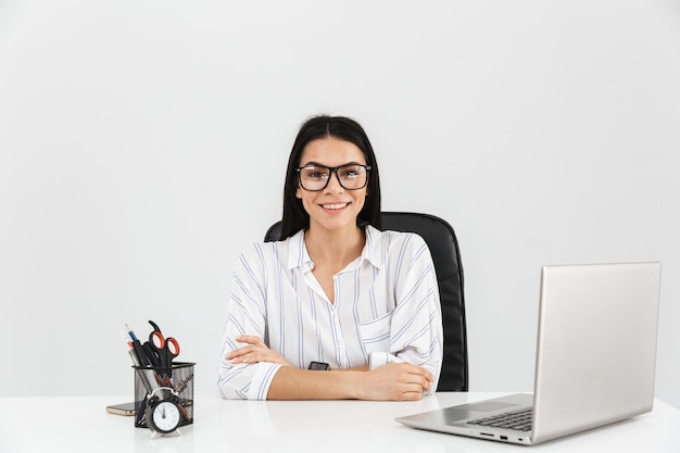 smart brunette businesswoman sitting at table avec papeterie et travaillant sur ordinateur portable au bureau isolé sur mur blanc