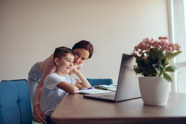 Smart gentil petit garçon élève apprenant en ligne sur un ordinateur portable et une mère étreignant