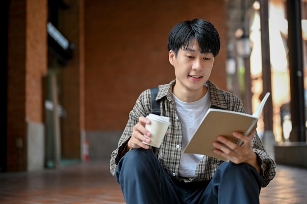 Smart young Asian male college student assis dans les escaliers à l'extérieur et lisant un livre