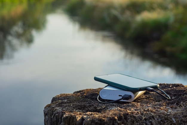 Le smartphone est chargé à l'aide d'un chargeur portable. Power Bank charge le téléphone à l'extérieur avec un sac à dos pour le tourisme en arrière-plan de la nature et de la rivière.