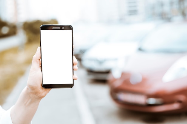 Smartphone à la main avec un écran blanc sur le fond des voitures en stationnement.