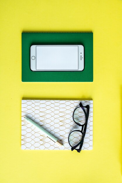 Smartphone sur ordinateur portable vert et stylo avec des lunettes sur un cahier sur fond jaune