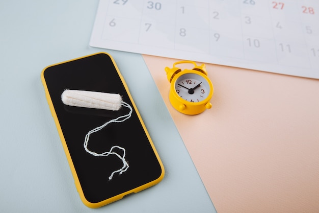 Smartphone Pour Suivre Votre Cycle Menstruel Et Pour Les Marques. Pms Et Le Concept Des Jours Critiques. Tampon En Coton, Tampon Quotidien Et Alarme Jaune Sur Fond Bleu Rose.