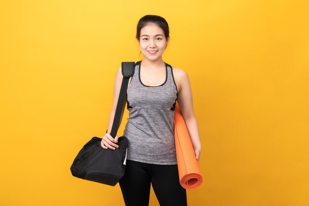 Smiley Asian woman wearing sportswear holding orange mat avec sac de sport sur le mur jaune Concept de mode de vie sain.