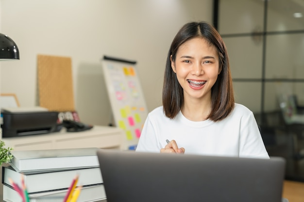 Smiley femme asiatique main tenant livre et stylo avec texte sous forme d'apprentissage sur ordinateur portable dans la maison.