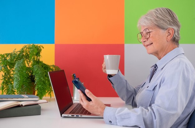 Photo smiling attractive senior woman holding a coffee cup et à l'aide de smart phone from office