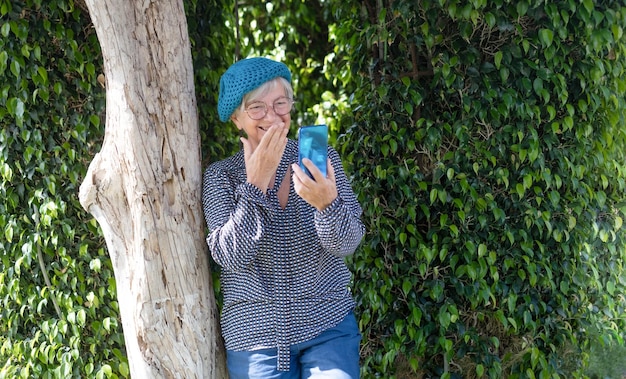 Smiling attractive senior woman sitting in the garden sous un arbre à l'aide de téléphone mobile en appel vidéo fond vert