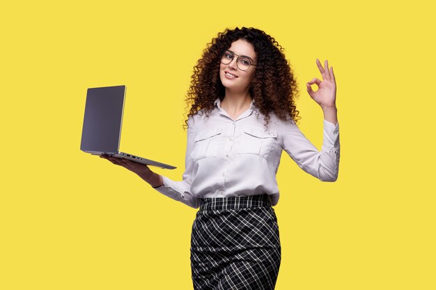 Smiling businesswoman on white shirt holding ordinateur portable et montrant ok geste du doigt isolé sur fond jaune