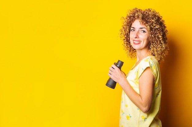 Smiling curly girl tenant des jumelles dans ses mains sur une surface jaune