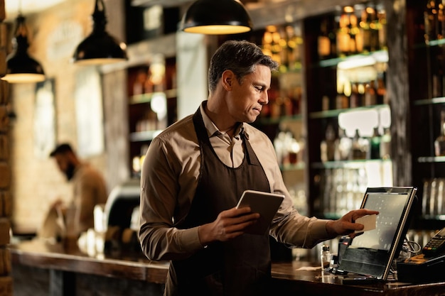 Smiling mid adult waiter using touchpad alors qu'il travaillait à la caisse enregistreuse dans un bar