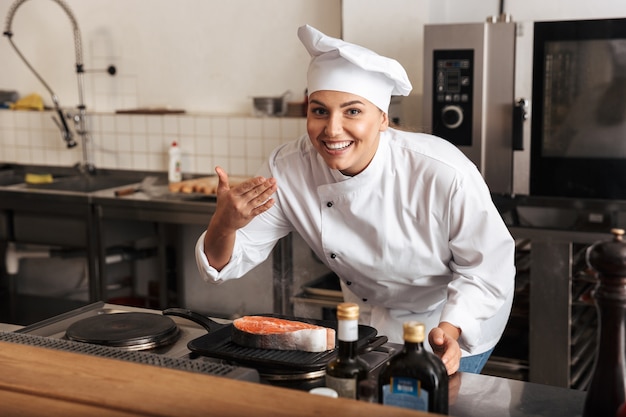 Smiling woman chef cuisinier en uniforme cuisson délicieux steak de saumon debout à la cuisine