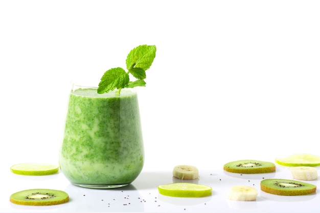 le smoothie vert se dresse sur une surface blanche avec des tranches de fruits à côté.