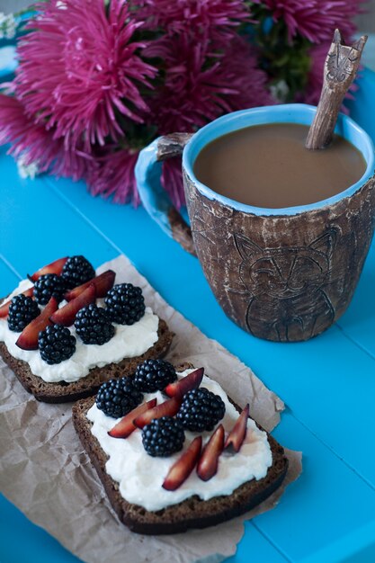 Sndwiches au fromage doux et aux baies, une tasse de café et un bouquet d'asters en surface en bois bleu