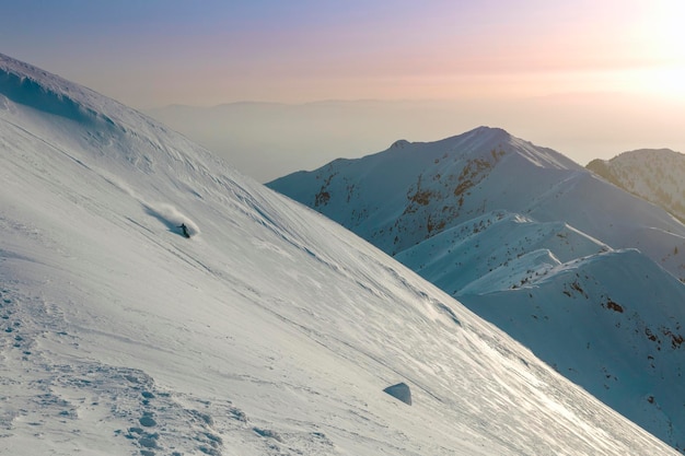 Un snowboarder descend une large pente sur fond de chaînes de montagnes et de coucher de soleil