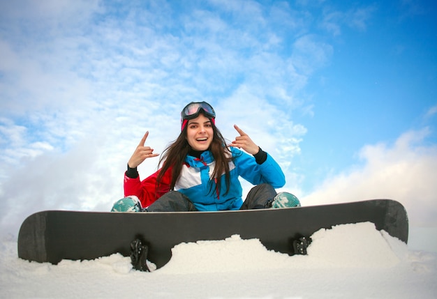 Snowboarder femme joyeuse assis au sommet de la montagne sur le ciel bleu