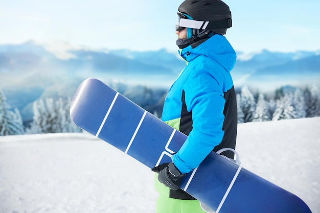 Snowboarder de l'homme à la station de ski sur l'arrière-plan Ciel bleu tenir snowboard portant des lunettes de ski lunettes de ski avec le reflet des montagnes enneigées
