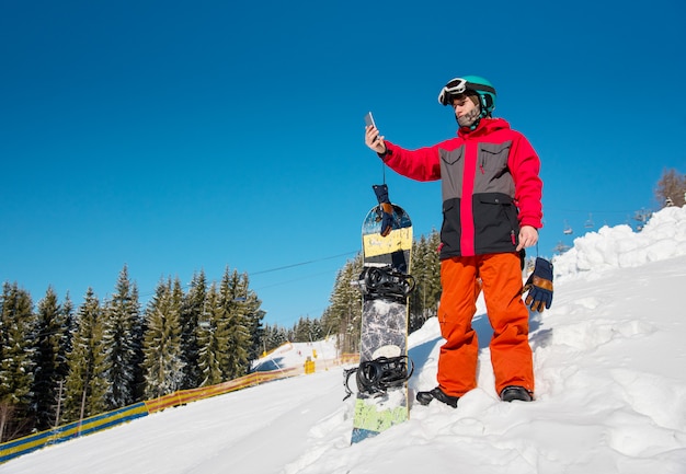 Snowboarder mâle reposant sur la pente de la station de ski, à l'aide de son téléphone intelligent après le snowboard