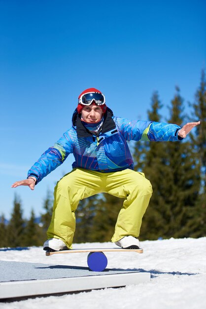 Snowboarder masculin en équilibre sur le snowboard dans la neige blanche sur fond de ciel bleu et d'épinettes