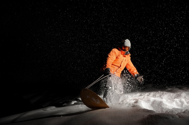 Snowboarder en orange sportswear et masque à cheval sur une colline enneigée la nuit