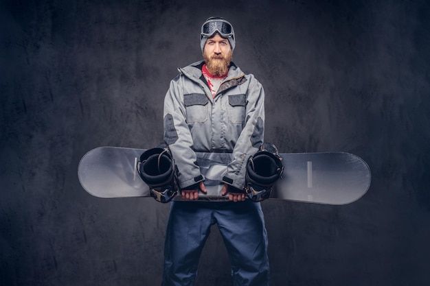 Snowboarder rousse brutal avec une barbe pleine dans un chapeau d'hiver et des lunettes de protection vêtu d'un manteau de snowboard tient un snowboard dans un studio. Isolé sur un fond texturé sombre.