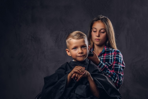 La sœur aînée coupe son petit frère avec une tondeuse sur le fond sombre. Mignon garçon d'âge préscolaire se coupe les cheveux.