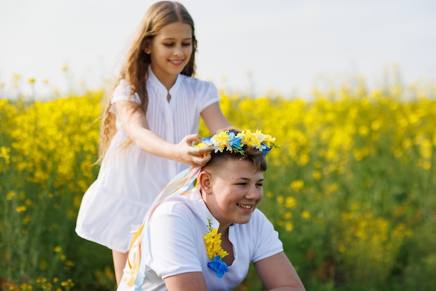 Sœur tresse des rubans dans une couronne ukrainienne avec des fleurs sur la tête du frère aîné sur le pré contre le champ