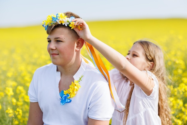 Sœur tresse des rubans dans une couronne ukrainienne sur la tête de son frère adolescent sur fond de champs et de ciel