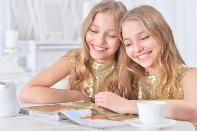 Sœurs jumelles mignonnes assises à la table de la cuisine avec un magazine moderne