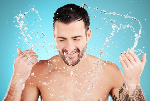 Soin de la peau à l'eau et homme avec éclaboussures faciales sourire et traitement de nettoyage du matin isolé sur fond bleu Hygiène du visage et modèle masculin toilettage pour la santé, le bien-être et les soins de la peau propre en studio