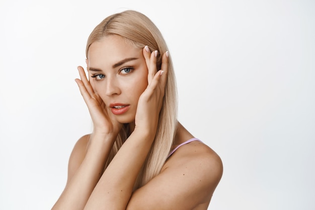 Soin de la peau. Portrait en gros plan d'une jolie femme blonde, touchant son visage près des yeux, semblant sensuel, concept de produits cosmétiques et spa sur blanc