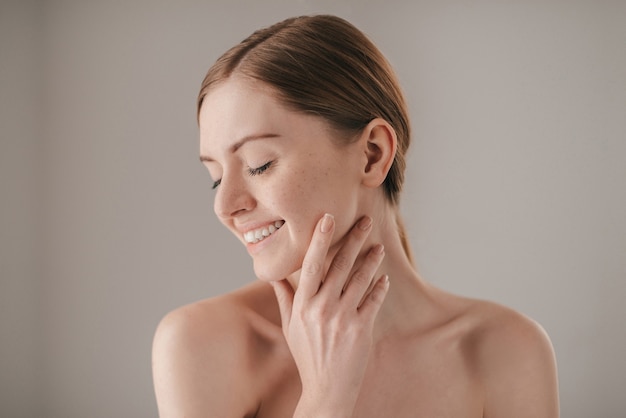 Soin sensible pour une peau lumineuse. Portrait de femme rousse avec des taches de rousseur gardant les yeux fermés et souriant tout en touchant son visage et debout sur fond gris