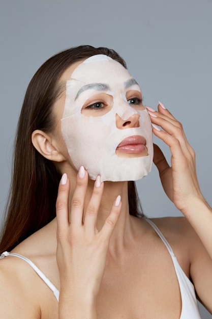 Soins de la peau du visage de beauté. La femme applique un masque hydratant en tissu sur le visage. Modèle de fille avec masque cosmétique. Traitement facial