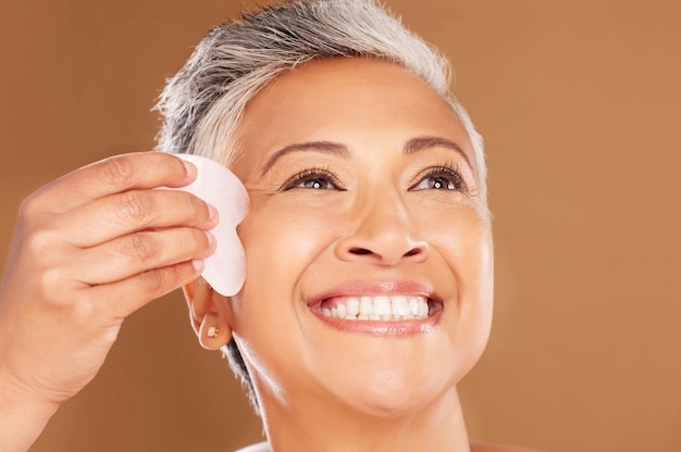 Photo soins de la peau gua sha et femme âgée avec un massage pour le bien-être du visage et un produit de luxe sur fond de studio de maquette marron vision de spa et modèle pensant à la beauté du visage avec un outil